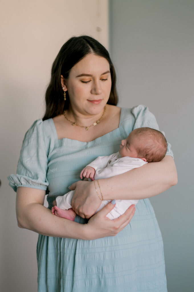 Cleveland In Home Newborn Photography Session by Brittany Serowski Photography. A new mother, in a light blue dress. Standing in her newborn son's nursery that is light blue and white themed. She is holding her newborn son cradled in her arms while looking down at him. The baby boy is sleeping and in all white romper.