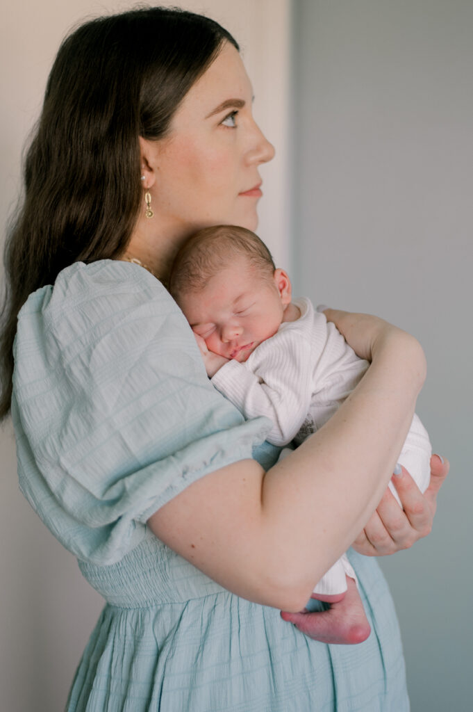 A new mother, holding her baby boy in his nursery. Baby is laying on her chest as she looks off into the distance. Mom is in a light blue dress and baby boy is in a white newborn romper. Photographed by Cleveland In Home Newborn Photography, Brittany Serowski Photography.