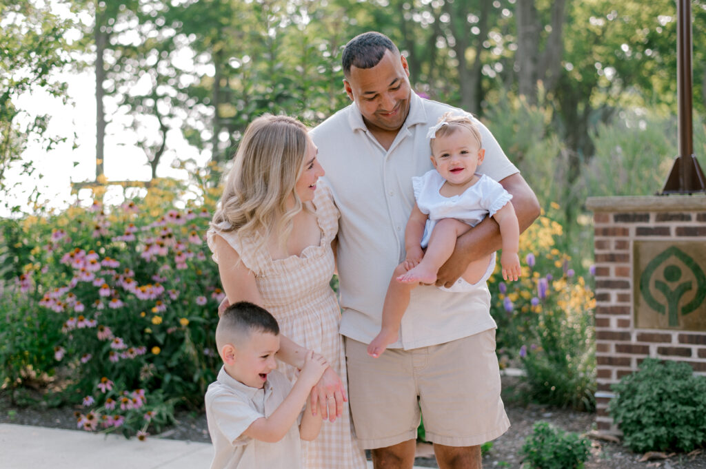 A family of four, all dressed in cream colors, stand together laughing as they are surrounded by the flowering gardens at Bay Village Huntington Beach Playground. Captured by Cleveland Family Photographer, Brittany Serowski Photography.