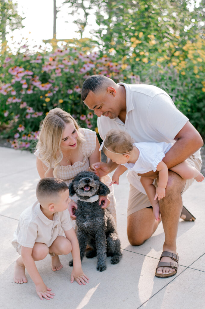 A family of four with their furry family member, standing in front of the flowering garden beds at Bay Village Huntington Beach Playground. Photographed by Cleveland Family Photographer, Brittany Serowski Photography.