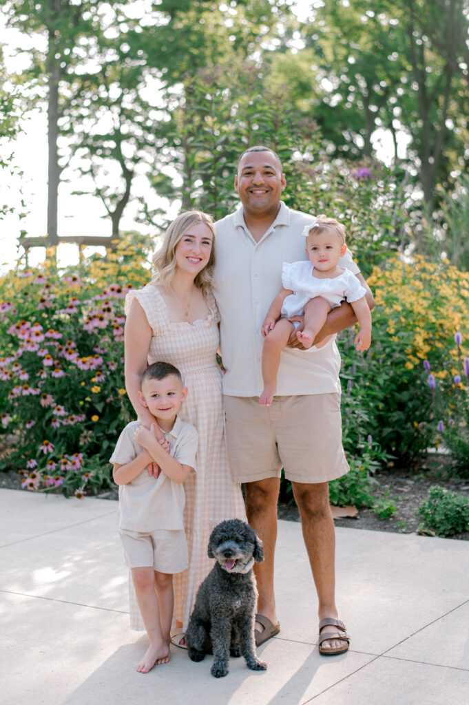 A young family standing and smiling at the camera with the flowering garden beds at Huntington Beach Playground in Bay Village. Photographed by Cleveland Family Photographer, Brittany Serowski Photography.