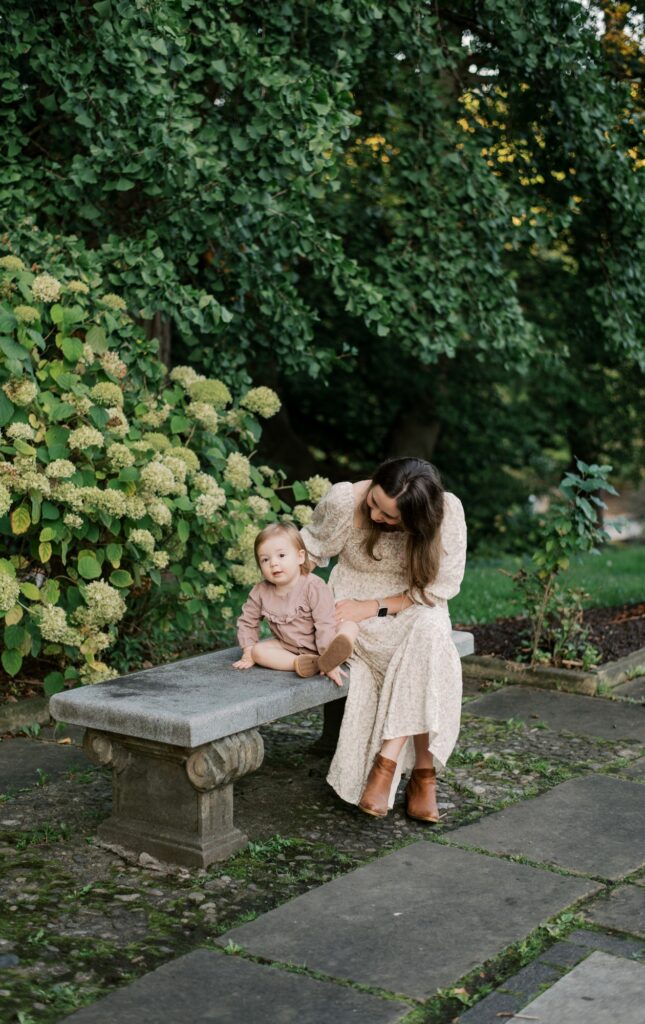 Spring flowers in Cleveland. A mother with her toddler daughter, sitting on a stone bench at the Cleveland Cultural Gardens. Behind them is lush trees, greenery and flowering hydrangeas. Captured by Cleveland Family Photographer, Brittany Serowski Photography.