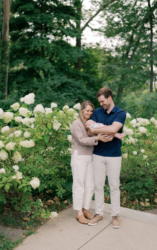 A mother and father, cradling their newborn son as they snuggle together in front of the flowering hydrangeas at the Cleveland Cultural Gardens for spring family photos in Cleveland. Photographed by Cleveland Family Photographer, Brittany Serowski Photography.